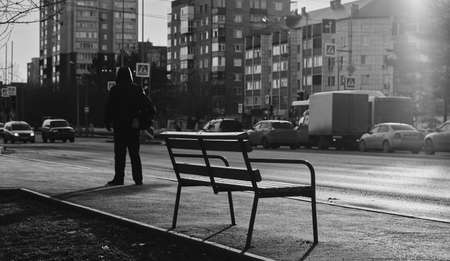 Black white photo of a man who is waiting for a bus standing next to an empty benchの写真素材