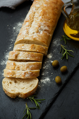 Ciabatta bread sliced, olives, olive oil and flour on a black stone surface with rosemary.の写真素材