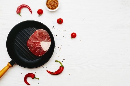Raw meat shank  with  dijon mustard, chili peppers and tomatoes in a grill frying pan on a white painted wooden surface. Top view. Flat lay. Copy space.の写真素材