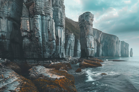 A dramatic coastal scene featuring towering cliffs carved by erosion, with the ocean waves crashing at their base against a backdrop of a cloudy sky.の素材
