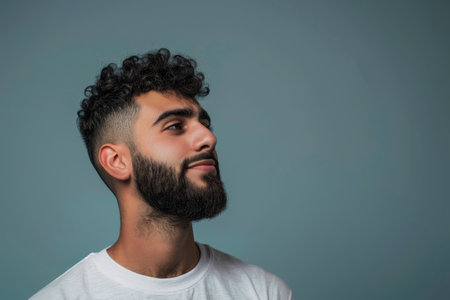 A portrait of a young man with curly hair and a beard looking to the side against a blue background.の素材