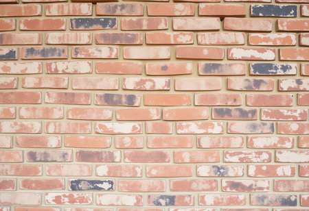 A close-up photograph of a weathered brick wall with a running bond pattern. The bricks are red, orange, brown, grey, and black, and they are arranged in horizontal rows. The mortar is beige and cream. The wall has a rough, uneven texture.の素材