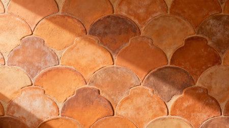 Overlapping terracotta fish-scale tiles covering an exterior wall, featuring varied reddish-brown textures and sharp diagonal sunlight casting dramatic relief shadows across the geometric design.の素材