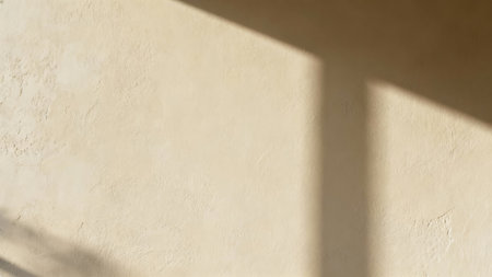Detailed view of rough, sandy colored plaster texture illuminated by directional window light, casting sharp, intersecting geometric shadows diagonally across the surface.の素材