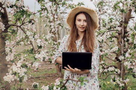 Girl in a straw hat and dress in the garden. Woman with a laptop and a flowering tree in the garden. Portrait.の写真素材