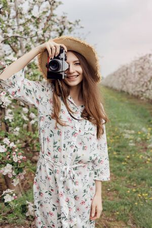 Nature Photographer taking pictures outdoors. Beautiful girl in a hat and dress in an apple orchard. Open air outside the city. Stylish woman between flowering trees with a camera.の写真素材