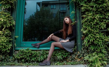 Adorable, young woman or cute girl standing near beautiful, blossoming flower pots on windows, floral decoration in street, on sunny, summer day on yellow house wall. Gardening. Flourishing, growingの写真素材