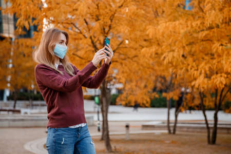 Girl with a phone in a mask. Woman in medical mask outdoors in autumn. Selfie in a mask.