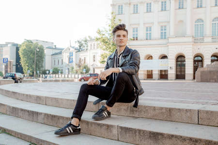 Attractive young hipster man in black jacket. walks in the city. Attractive guy on walk. Urban fresh portrait confident young man with hairstyle in fashion black denim jacket near wall outdoors.の写真素材