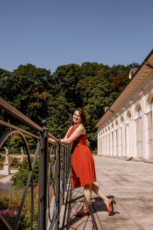 Beautiful girl in red dress posing near the white house. Portrait of beautiful young brunette muse woman. Portrait of pretty girl on the street of town. Walk in downtown. Palace in the Royal Lazienki. Warsaw.の写真素材