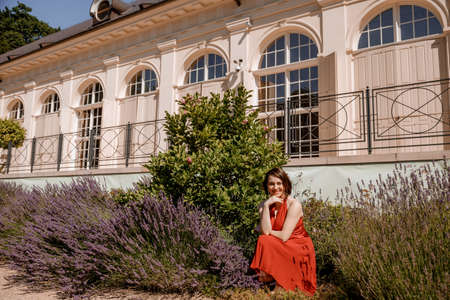 The Great Orangery a classicistic orangery in Lazienki Krolewskie park in Warsaw. Woman in Garden, Old White Alcove in Flowers, Outdoor Beauty Portrait. A woman with red dress sitting. Poland.の写真素材