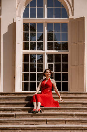 The Great Orangery a classicistic orangery in Lazienki Krolewskie park in Warsaw. Woman in Garden, Old White Alcove in Flowers, Outdoor Beauty Portrait. A woman with red dress sitting. Poland.の写真素材