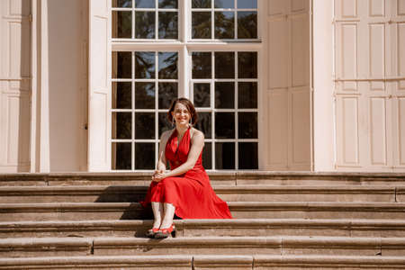 The Great Orangery a classicistic orangery in Lazienki Krolewskie park in Warsaw. Woman in Garden, Old White Alcove in Flowers, Outdoor Beauty Portrait. A woman with red dress sitting. Poland.の写真素材