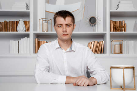 A man in a white shirt is sitting at a white table. Young business man on a desk. Attractive businessman sitting at table. Front view of a business man working.の写真素材