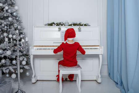 Boy in santa costume. Kid concept for merry Christmas and happy new year celebration. Small boy enjoys playing piano for the first time. Child play piano near the christmas tree. Christmas time.の写真素材