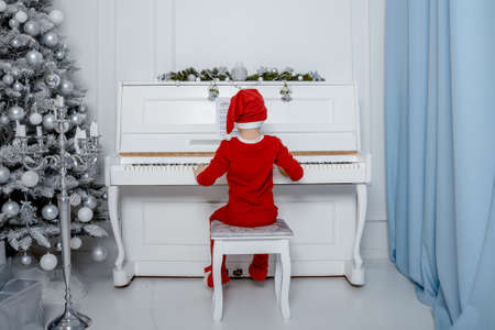 Boy in santa costume. Kid concept for merry Christmas and happy new year celebration. Small boy enjoys playing piano for the first time. Child play piano near the christmas tree. Christmas time.の写真素材