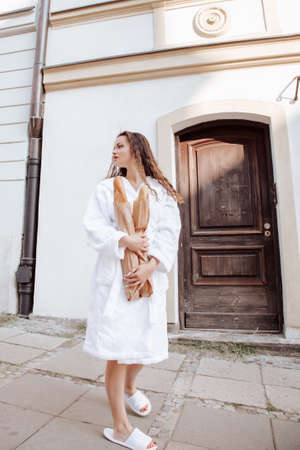 Young woman in white robe looks at street of city. Attractive girl holding baguettes and paper bag with purchases. Young stylish woman buying a french baguette standing on the street in Warsaw city.の写真素材