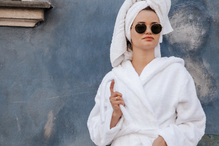 Smiling cute girl blogger in white robe, towel and sunglasses stands on old street. Portrait young...