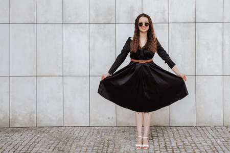 Stylish girl in a black dress and sunglasses near the gray wall. Photo of woman in glasses. Young woman, curly hairstyle, smiling near a wall in the street. Girl wearing black dress clothes.の写真素材