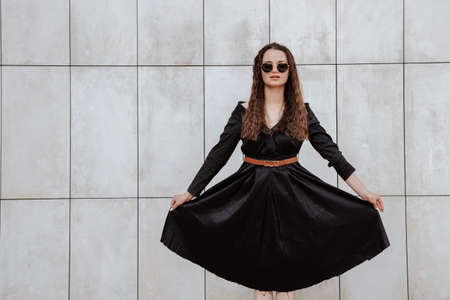 Stylish girl in a black dress and sunglasses near the gray wall. Photo of woman in glasses. Young woman, curly hairstyle, smiling near a wall in the street. Girl wearing black dress clothes.の写真素材