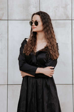 Stylish girl in a black dress and sunglasses near the gray wall. Photo of woman in glasses. Young woman, curly hairstyle, smiling near a wall in the street. Girl wearing black dress clothes.の写真素材