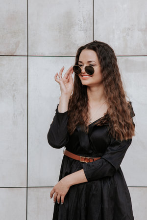 Stylish girl in a black dress and sunglasses near the gray wall. Photo of woman in glasses. Young woman, curly hairstyle, smiling near a wall in the street. Girl wearing black dress clothes.の写真素材