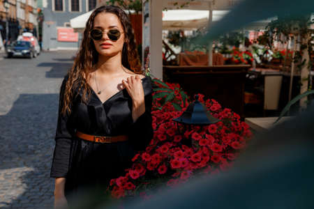 Cute stylish model in sunglasses posing near the flower bush on the city street. Beautiful young woman with long hair, wearing black dress, posing near flowering bushes on the old city street. Warsaw.の写真素材