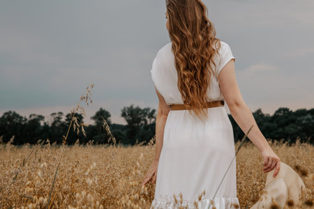 Beautiful young girl dressed in a white dress and hat walks in the field and poses of oats at...