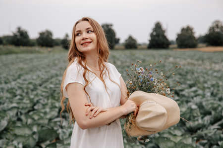 A beautiful slender girl with a bouquet of wildflowers on the background of on cabbage field. Woman...