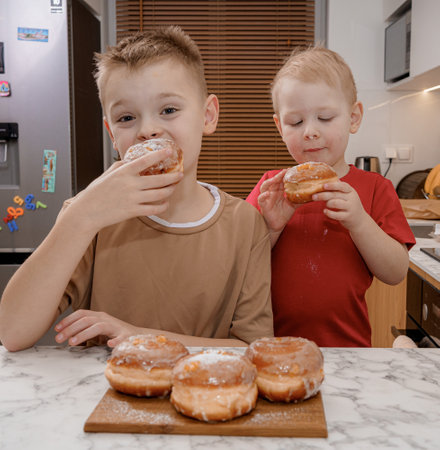 Two young boys eagerly admires a stack of traditional Polish packs, glazed and filled with custard, on a kitchen counter. They sniffs the sweet treat, symbolizing the joy of Fat Thursday indulgence.の写真素材