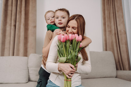 A woman sits on a sofa holding a bouquet of pink tulips, embraced by her two smiling children. A heartwarming scene full of love and family bonding, perfect for International Women's Day.の写真素材