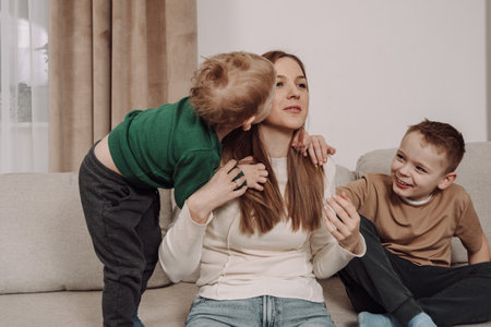 A happy mother embraces her children in a warm, homey atmosphere. The younger boy kisses her cheek, while the older one smiles joyfully. A natural, heartfelt family moment full of love.の写真素材