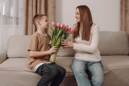 A sweet boy hugs his mother and gives her a bouquet of pink tulips. A happy mother and her son share a tender, loving moment in the comfort of their home. A perfect image for Mother's Day, International Women's Day, and campaigns related to family, motherhood, and gratitude.の写真素材
