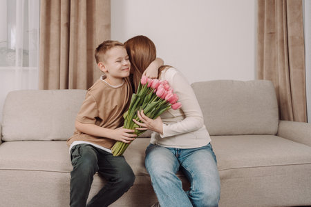 A sweet boy hugs his mother and gives her a bouquet of pink tulips. A happy mother and her son share a tender, loving moment in the comfort of their home. A perfect image for Mother's Day, International Women's Day, and campaigns related to family, motherhood, and gratitude.の写真素材