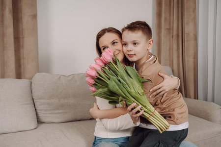 A sweet boy hugs his mother and gives her a bouquet of pink tulips. A happy mother and her son share a tender, loving moment in the comfort of their home. A perfect image for Mother's Day, International Women's Day, and campaigns related to family, motherhood, and gratitude.の写真素材