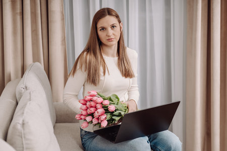 Young woman in casual outfit sitting on a couch with a laptop, looking away. Pink tulips beside her. Cozy home interior, work-from-home concept, modern lifestyle.の写真素材