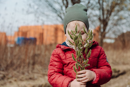 A young child in a red jacket and green hat holds a bouquet of fluffy willow branches outdoors in...