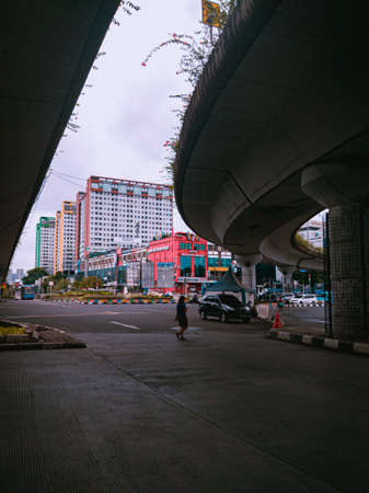 Jakarta Pusat, Indonesia - 14 February 2021: A woman in blue is crossing a highway that is located under two concrete bridges against a backdrop of colorful buildings.のeditorial素材