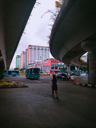 Jakarta Pusat, Indonesia - 14 February 2021: A woman in blue is crossing a highway that is located under two concrete bridges against a backdrop of colorful buildings.のeditorial素材