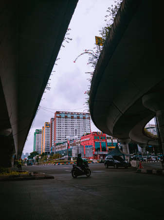 Jakarta Pusat, Indonesia - 14 February 2021: Motorbikes cross the highway located under two concrete bridges against a backdrop of colorful buildings.のeditorial素材