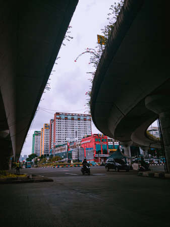 Jakarta Pusat, Indonesia - 14 February 2021: Motorbikes cross the highway located under two concrete bridges against a backdrop of colorful buildings.のeditorial素材
