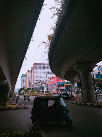 Jakarta Pusat, Indonesia - 14 February 2021:  Bajaj cars cross the highway which is located under two concrete bridges with a background of colorful buildings.のeditorial素材