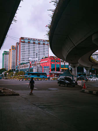 Jakarta Pusat, Indonesia - 14 February 2021: A woman in blue is crossing a highway that is located under two concrete bridges against a backdrop of colorful buildings.のeditorial素材