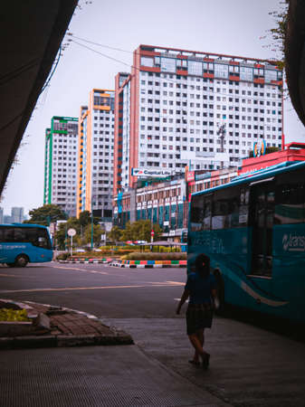 Jakarta Pusat, Indonesia - 14 February 2021: A woman in blue is crossing a highway that is located under two concrete bridges against a backdrop of colorful buildings.のeditorial素材