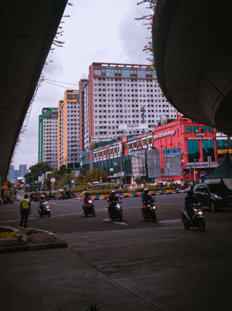 Jakarta Pusat, Indonesia - 14 February 2021: Motorbikes cross the highway located under two concrete bridges against a backdrop of colorful buildings.のeditorial素材