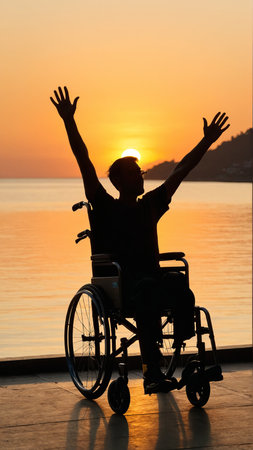 Silhouette of person in wheelchair celebrating at sunset by the sea.の素材