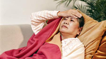Elderly woman resting with blanket feeling unwell on couch.の素材