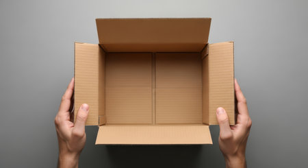 Hands holding open cardboard box on gray background for shipping and packaging conceptsの素材