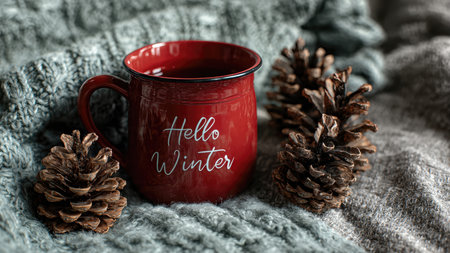 Cozy red mug with hello winter text surrounded by pine cones on knitted blanket.の素材