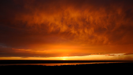 A magnificent sunset paints the sky in deep orange and red hues, with dramatic, textured clouds creating a fiery canopy above the horizon. The sun gently dips below the edge, illuminating the landscape and reflecting on the tranquil river below. The contrast between the dark silhouettes and glowing sky evokes a sense of awe and natural splendor.の素材